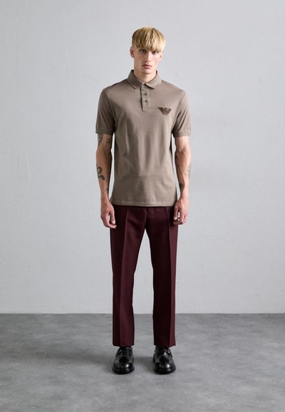 Polo marron avec un logo sur la poitrine gauche, associé à un pantalon bordeaux foncé et des chaussures noires. Texture en coton lisse et coupe décontractée.