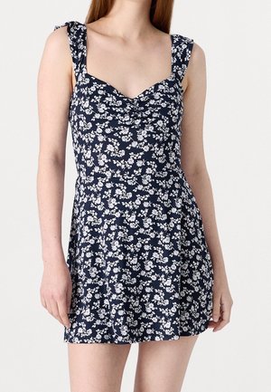 Žena v tmavě modrých minišatech bez rukávů s bílým květinovým potiskem a nařaseným živůtkem, stojící před jednobarevným světlým pozadím.