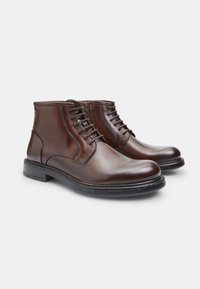 Botas de cuero marrón hasta el tobillo con una textura suave, que presentan un cierre de cordones y cremallera, punta redonda y un diseño de tacón apilado.