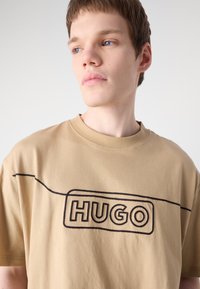 Beige bomull t-skjorte med svart brodert "HUGO" logo, rund halsdesign, korte ermer og en avslappet passform. Glatt tekstur.