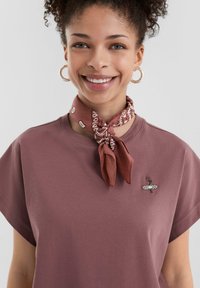 T-shirt bordeaux à manches courtes avec une pièce en tissu. Une écharpe à motifs dans des teintes chaudes est nouée autour du cou, ajoutant texture et détail.