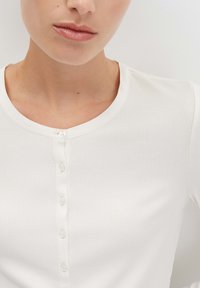 Close-up van een persoon in een witte geribbelde button-up shirt met een ronde hals, waarbij de lippen, kin en bovenlichaam zichtbaar zijn.