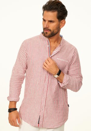 Mann trägt ein langärmeliges Hemd mit roten und weißen vertikalen Streifen, richtet den Kragen, trägt eine schwarze Uhr und Armbänder und schaut nach links.