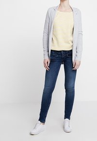 Cardigan grigio, top a righe gialle e bianche, jeans skinny blu scuro e scarpe da ginnastica bianche. Design minimalista senza motivi visibili né accessori.