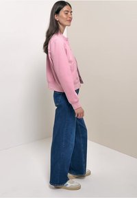 Lichtroze sweatshirt met geribde manchetten, gecombineerd met een brede blauwe denimbroek en beige sneakers met witte en blauwe accenten.