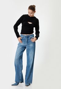 Schwarzer gerippter Strickpullover mit einem Ausschnitt, kombiniert mit hellblauen weit geschnittenen Denim-Jeans und blauen, spitzen Absatzmules.