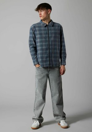 Pledēta josta zilā un pelēkā toņos, kas savienota ar gaiši pelēcīgām vaļīgām džinsi un baltām teniskām ar brūnganiem akcentiem.