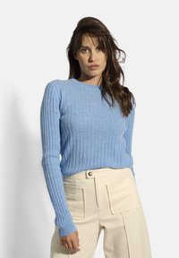 Maglione a coste blu cielo con maniche lunghe e scollatura rotonda, abbinato a pantaloni color crema con tasche frontali e dettaglio dei bottoni.