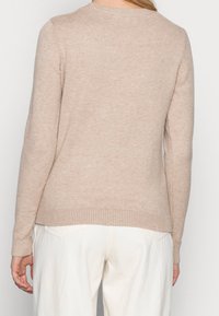 Beżowy sweter dzianinowy z okrągłym dekoltem, prążkowanymi mankietami i dołem. Miękka tekstura, klasyczny krój i prosty design. Noszony z jasnymi spodniami.
