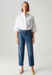 Camicia bianca con bottoni e una tasca sul petto, abbinata a jeans blu a vita media e gamba dritta e scarpe beige senza lacci. Design minimalista.