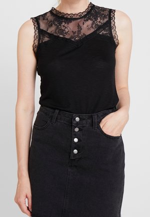 Musta toppi, jossa on pitsityyppinen koriste kaula-aukon ja olkapäiden alueella, yhdistetty korkeavyötäröiseen mustaan farkkuhameeseen, jossa on hopeanväriset napit.