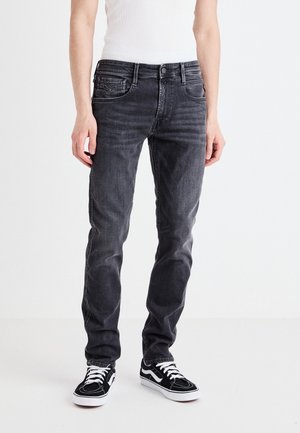 Persona con jeans neri slim-fit scoloriti, canottiera bianca e sneakers bianche e nere in piedi su sfondo semplice.