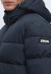 Homme portant une veste matelassée bleu marine foncé avec capuche et patch logo Schott New York sur le haut de la manche.