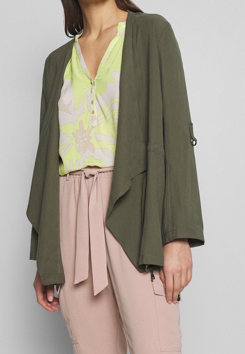 Femme portant une veste ouverte vert olive par-dessus un chemisier floral vert clair et un pantalon taille haute rose clair avec une ceinture nouée.