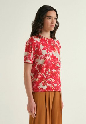 Donna che indossa un top a maniche corte floreale rosso e beige e una gonna plissettata marrone, in piedi davanti a uno sfondo chiaro uniforme.