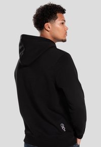 Sudadera con capucha negra, mezcla de algodón, que presenta un pequeño diseño bordado en la parte inferior de la espalda. Textura suave y corte relajado.