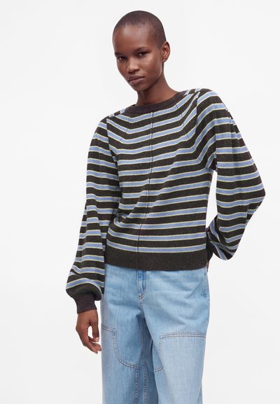 Personne portant un pull sombre à rayures avec des lignes bleues et jaunes et un jean bleu clair à poches plaquées, debout avec une main sur la hanche.