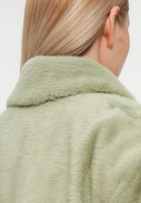 Zelený kožich z umělé kožešiny s klopovým límcem, měkkou texturou a mírně oversized střih. Zaměření je na zadní a ramenní část.