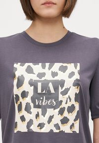 Siva majica kratkih rukava s velikim grafičkim uzorkom leopard tiska u zlatnim akcentima i tekstom "LA vibes" u središtu.