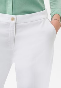 I pantaloni bianchi presentano un frontale piatto con una chiusura a un solo bottone, tasche laterali e un tessuto liscio e leggero. La vita è ampia.