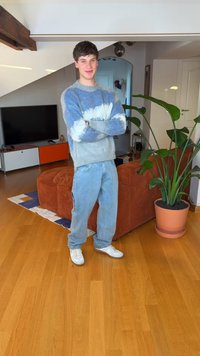 Trui in blauwgradiënt en grijs, loszittende spijkerbroek, witte sneakers, staand in een kamer met houten vloeren en een plant.