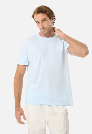 Homme aux cheveux bouclés bruns portant un T-shirt bleu clair et un pantalon blanc, touchant l'arrière de son cou avec la main droite, debout devant un fond blanc.