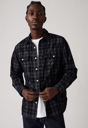 Black and white plaid button-up shirt with two chest pockets, long sleeves, and white buttons, layered over a white crew neck T-shirt.