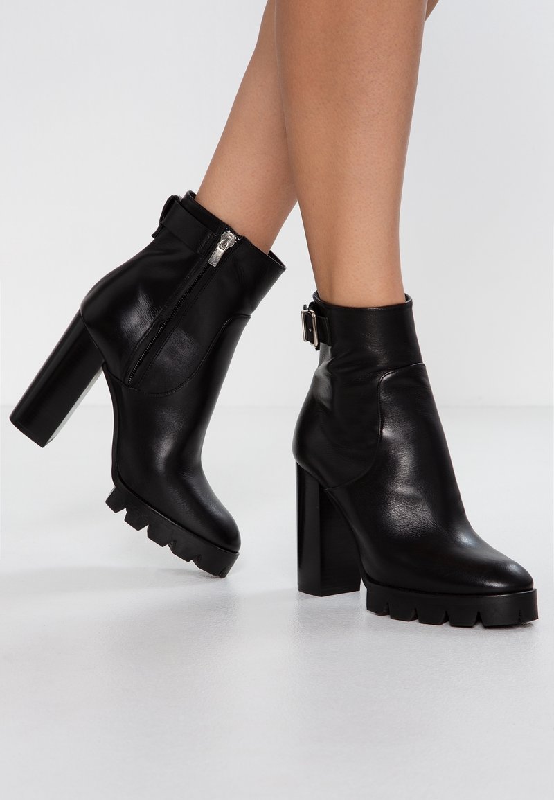 Bottes courtes en cuir noir avec un talon haut à bloc, une fermeture éclair latérale et un détail de sangle à la cheville. Semelle épaisse avec des crampons prononcés. Texture lisse.