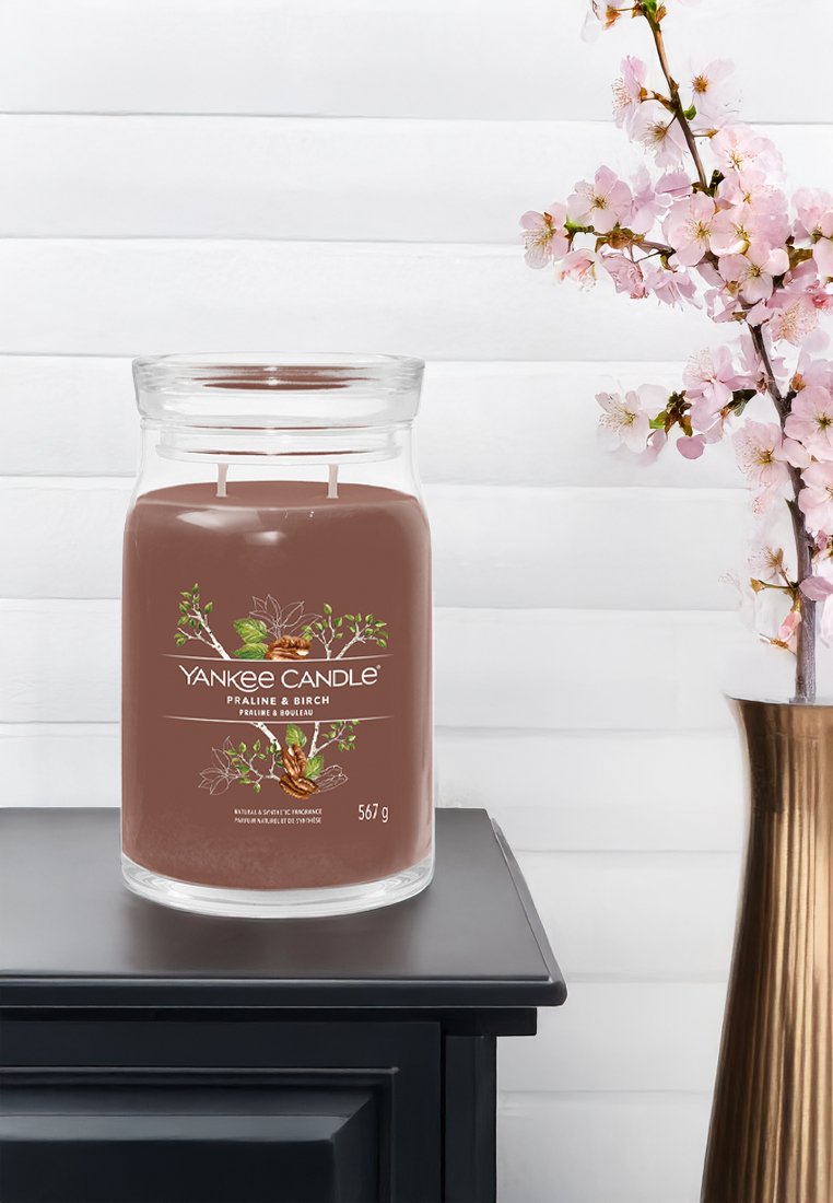 Bougie Yankee Candle avec une cire brune et deux mèches. Pot en verre avec l'étiquette "Praliné & Bouleau", posé sur une surface sombre à côté de fleurs roses.