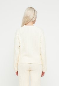 Pulover alb cu model împletit, cu croială lejeră, mâneci lungi și decolteu rotund, asortat cu pantaloni texturati asortați.