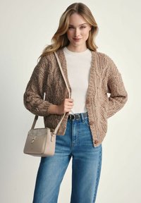 Kobieta nosząca brązowy sweter o grubym splocie, białą koszulę, niebieskie dżinsy, czarny pasek z złotymi pierścionkami i beżową torebkę na ramię, uśmiechająca się do kamery.