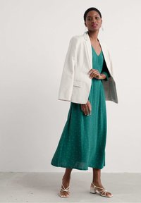 Vestido verde hasta la rodilla con lunares blancos, combinado con una chaqueta beige de gran tamaño y tacones de tiras de color claro. Tela suave, corte holgado.