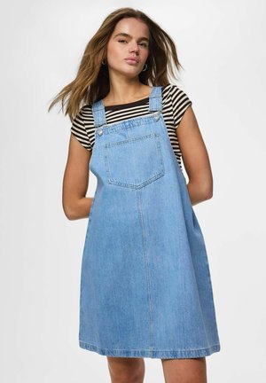 Ung kvinde med løst brunt hår iført en stribet skjorte under en lyseblå denimspencer kjole mod en ensfarvet baggrund.