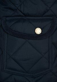 Tascone in tessuto blu scuro trapuntato con pattina curva chiusa da un bottone tondo dorato emblema di lettere.