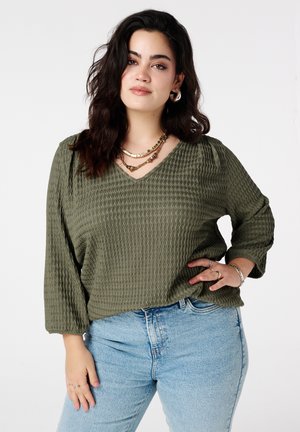 Vrouw met lang donker haar die een gestructureerde olijfgroene top, gelaagde gouden kettingen, lichtgewassen spijkerbroek en zilveren sieraden draagt, poserend met één hand op de heup.