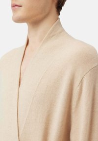 Beige stickad kofta med bred sjalkrage. Tyget verkar mjukt med en slät yta. Minimalistisk design med en avslappnad passform.