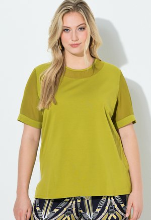 Femme aux longs cheveux blonds portant un haut vert citron à manches courtes et un pantalon à motifs noirs, blancs et jaunes, debout contre un mur clair.