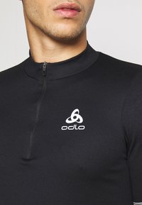 Černé sportovní tričko s dlouhým rukávem, s polovičním zipem a texturovaným materiálem. Na přední straně se nachází bílé logo Odlo.