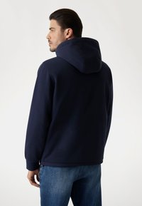 Felpa navy con cappuccio a contrasto, realizzata in tessuto testurizzato. Presenta polsini e orlo a coste, e dettagli con zip laterale. Indossata con jeans blu.