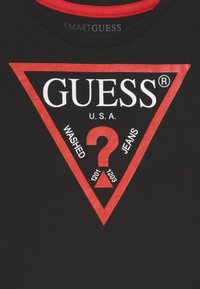 Mustapaita, jossa on punainen kolmio-logo. Mukana teksti "GUESS® U.S.A." sekä kysymysmerkki ja numerot 1201 ja 1203. Punainen kaulus.