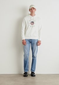 Hvit hettegenser med logo og rød, blå sirkel, kombinert med lyseblå denimjeans og svarte sko. Modell som bærer en beige caps.