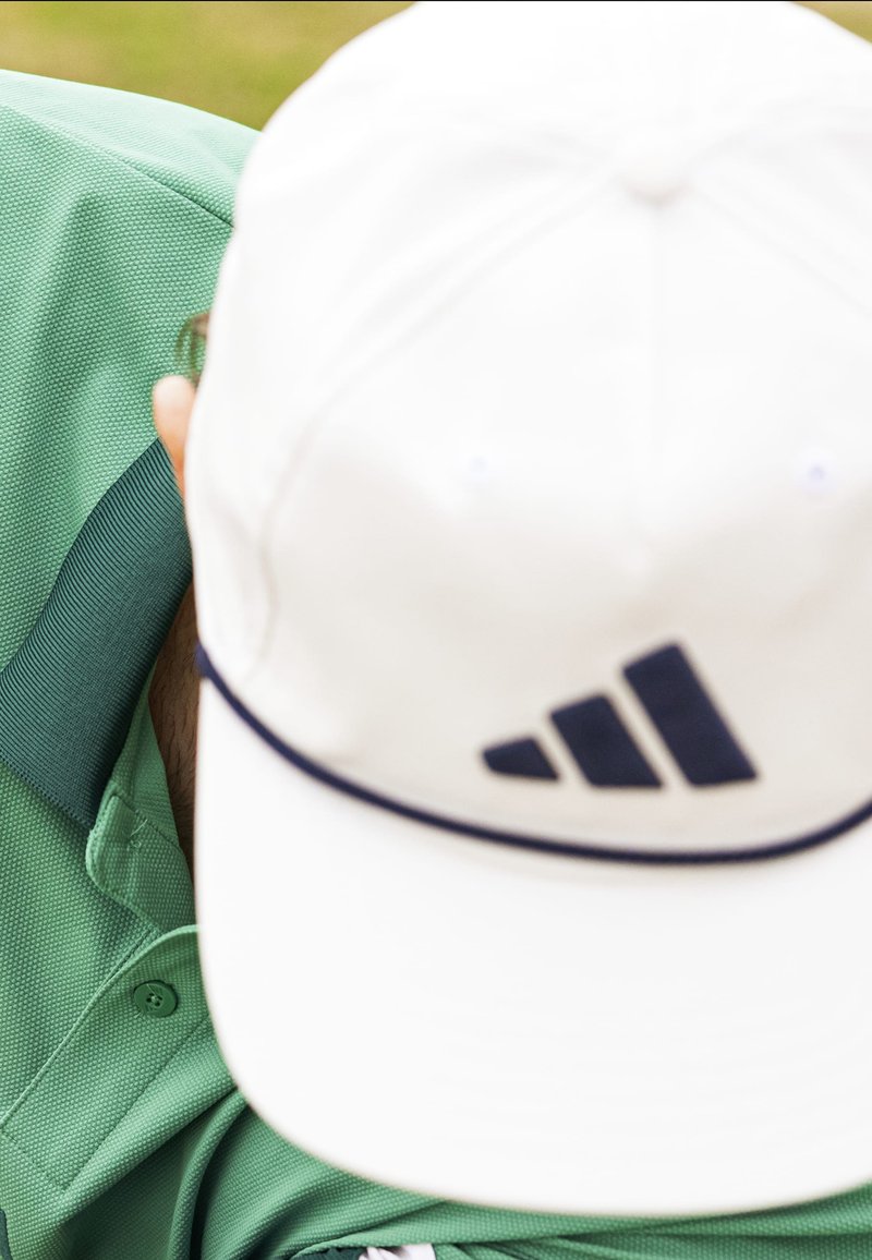 Casquette de baseball blanche avec logo bleu marine, portée légèrement de travers, associée à un polo vert texturé avec un détail de bouton et un col contrastant.