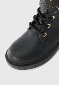 Schwarze Lederstiefel mit runder Spitze, ausgestattet mit goldenen Ösen und schwarzen Schnürsenkeln. Dekoratives, durchbrochenes Blumenmuster am Knöchel.