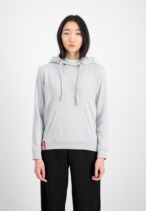 Young woman wearing a light gray hoodie with front pocket and drawstrings, paired with black pants, standing against a white background.