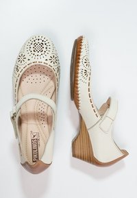 Valkoiset nahkakengät, joissa on rei'itetyt kukkakuviot, suljettu kärki, takasanka ja ruskea vaahtopohja, jossa on pystysuorat raidat.