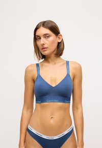 Žena s kratkom smeškom kestenjavom kosom koja nosi plavi Calvin Klein grudnjak i odgovarajuće plave gaće s bijelim pojasom na jednobojnoj pozadini.