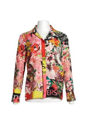 Camisa de manga larga con botones y patrones florales multicolor vibrantes y elementos de texto mixtos en un torso de maniquí blanco.
