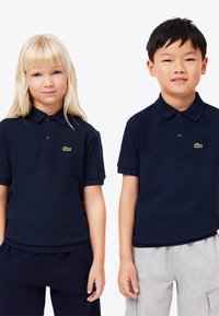 Blondin tyttö ja mustahiuksinen poika seisovat vierekkäin navy-sinisiä polo-paitoja päällään, joissa on vihreä krokotiililogo; tyttö pitää navy-sinisiä housuja, poika harmaita housuja.