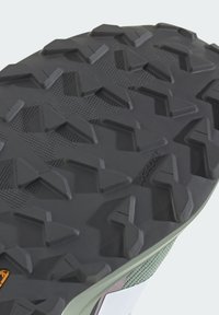 Semelle en caoutchouc noir avec un motif de crantage profond et angulaire et une texture antidérapante. Présente un dessus en maille verte avec des accents contrastants.