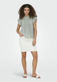 Lichtgroene blouse met kantpatroon en ruches aan de mouwen, gecombineerd met een witte denim rok en lichte sandalen. Vlotte zomeroutfit.